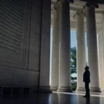 Homem de terno, enquadrado à grandiosidade de um monumento de mármore com escritos em inglês. Imagem do documentário "The Age of Disclosure".