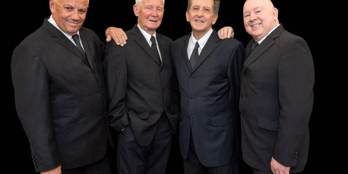 Foto dos quatro membros da banda "The Searchers" vestindo ternos pretos e sorrindo posando para a foto. Os dois membros das extremidades põe as mãos nos ombros nos dos do meio. O fundo da foto é preto.