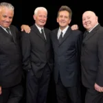 Foto dos quatro membros da banda "The Searchers" vestindo ternos pretos e sorrindo posando para a foto. Os dois membros das extremidades põe as mãos nos ombros nos dos do meio. O fundo da foto é preto.