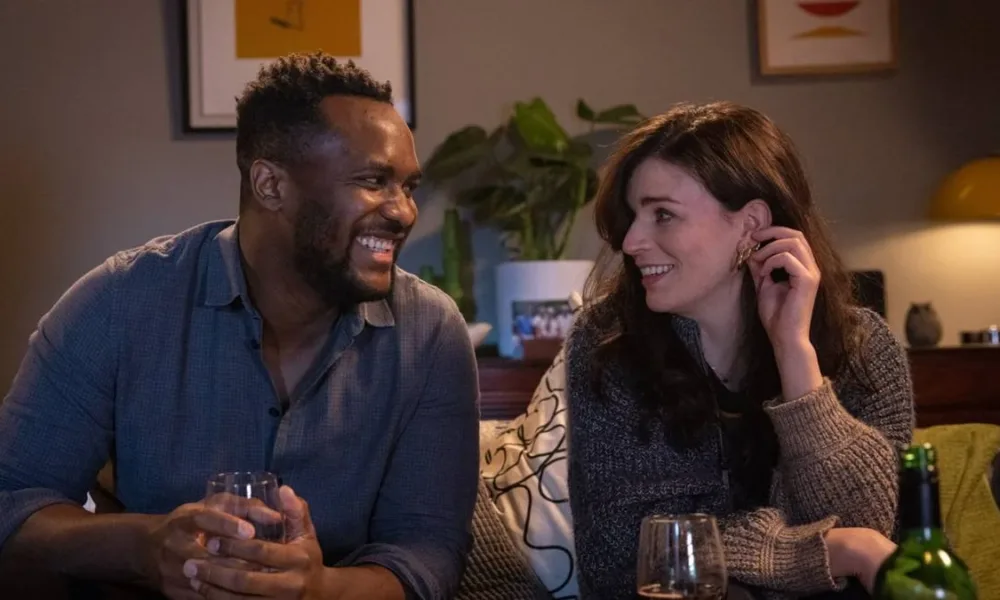 Kadiff Kirwan, esquerda, e Aisling Bea, direita, trocando olhares e sorrindo enquanto tomam vinho. Eles interpretam seus personagens na série "This Way Up". E estão sentados em um sofá.