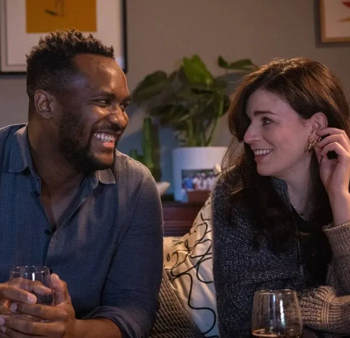 Kadiff Kirwan, esquerda, e Aisling Bea, direita, trocando olhares e sorrindo enquanto tomam vinho. Eles interpretam seus personagens na série "This Way Up". E estão sentados em um sofá.