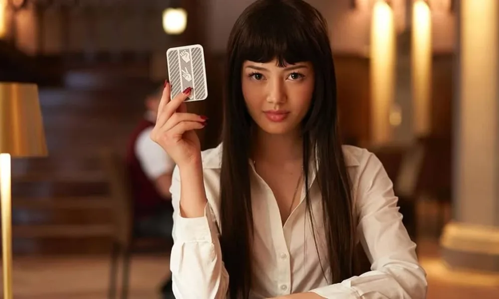 Protagonista de 'BET', adaptação live-action de "Kakegurui". Protagonista está com a mão esquerda na mesa, cotovelo direito apoiado e segurando uma carta com a mão direita, encarando o espectador, vestindo uma camisa branca.