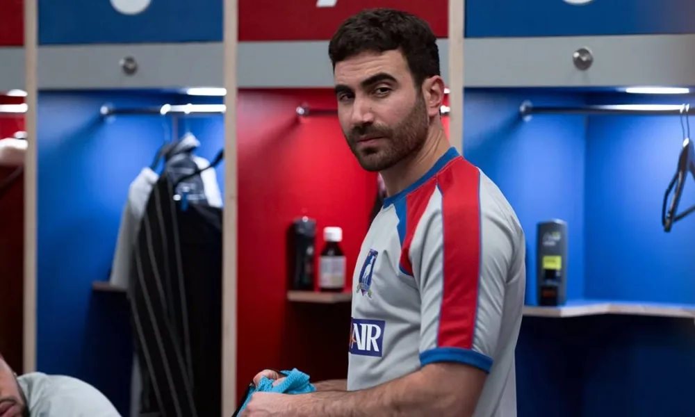 Brett Goldstein no episódio 9 da primeira temporada de "Ted Lasso" interpretando Roy Kent. Ele está num vestiário, com camisa de time, e um olhar dissimulado.