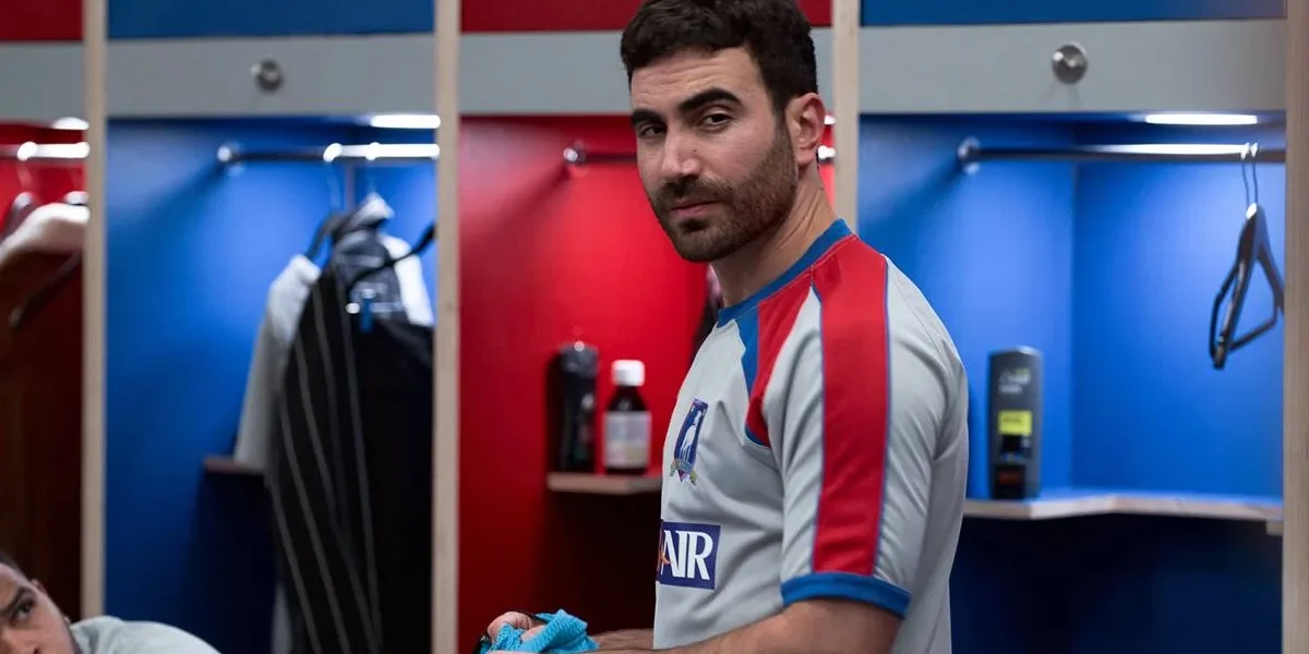 Ted Lasso | Brett Goldstein Faz Piada Com o Retorno da Série Brett Goldstein no episódio 9 da primeira temporada de "Ted Lasso" interpretando Roy Kent. Ele está num vestiário, com camisa de time, e um olhar dissimulado.