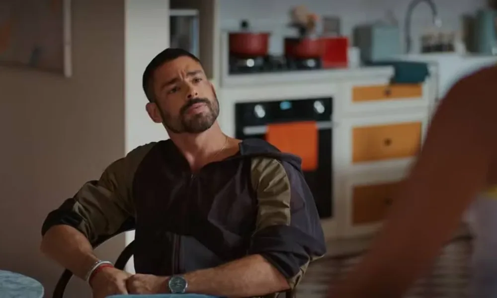 Cauã Reymond como César Ribeiro em "Vale Tudo". Personagem está segurando as próprias mãos, sentado em uma mesa, de relógio, olhando para Maria de Fátima, de costas para a câmera, interpretada por Bella Campos.