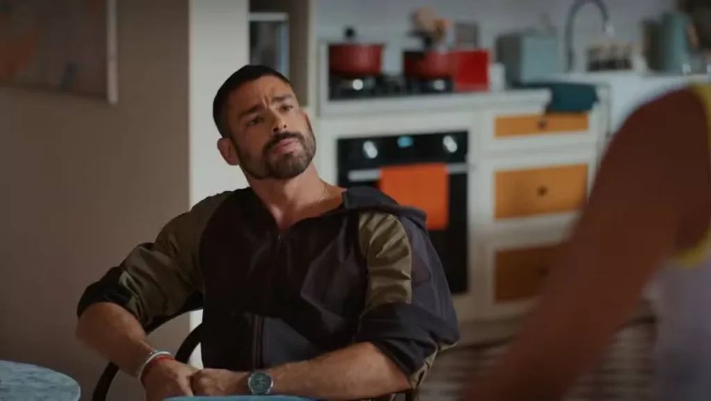Cauã Reymond como César Ribeiro em "Vale Tudo". Personagem está segurando as próprias mãos, sentado em uma mesa, de relógio, olhando para Maria de Fátima, de costas para a câmera, interpretada por Bella Campos.
