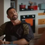 Cauã Reymond como César Ribeiro em "Vale Tudo". Personagem está segurando as próprias mãos, sentado em uma mesa, de relógio, olhando para Maria de Fátima, de costas para a câmera, interpretada por Bella Campos.