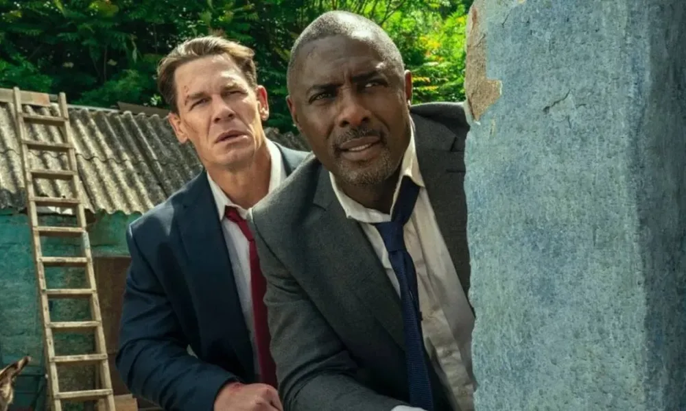 John Cena (esquerda e atrás) e Idris Elba (direita e frente), escondidos atrás de um muro vestindo ternos na comédia "Chefes de Estado".
