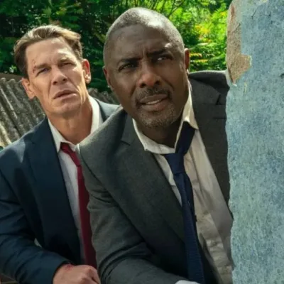 John Cena (esquerda e atrás) e Idris Elba (direita e frente), escondidos atrás de um muro vestindo ternos na comédia "Chefes de Estado".
