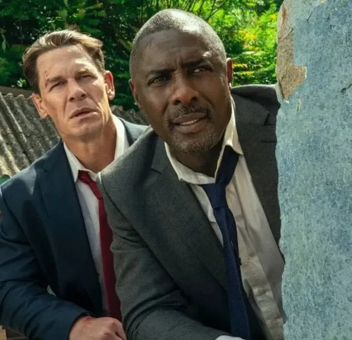 John Cena (esquerda e atrás) e Idris Elba (direita e frente), escondidos atrás de um muro vestindo ternos na comédia "Chefes de Estado".