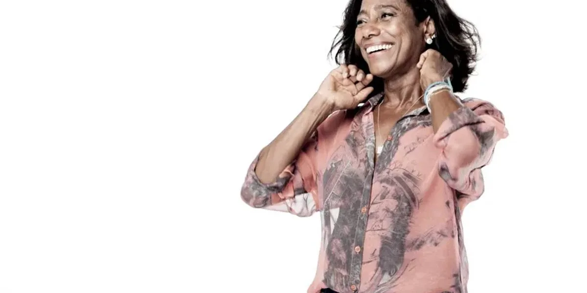 Gloria Maria em um fundo branco, sorrindo, sendo vista pouco abaixo da cintura para cima, mãos na altura das têmporas meio-fechadas, e com um sorriso no rosto.