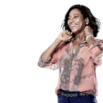 Gloria Maria em um fundo branco, sorrindo, sendo vista pouco abaixo da cintura para cima, mãos na altura das têmporas meio-fechadas, e com um sorriso no rosto.