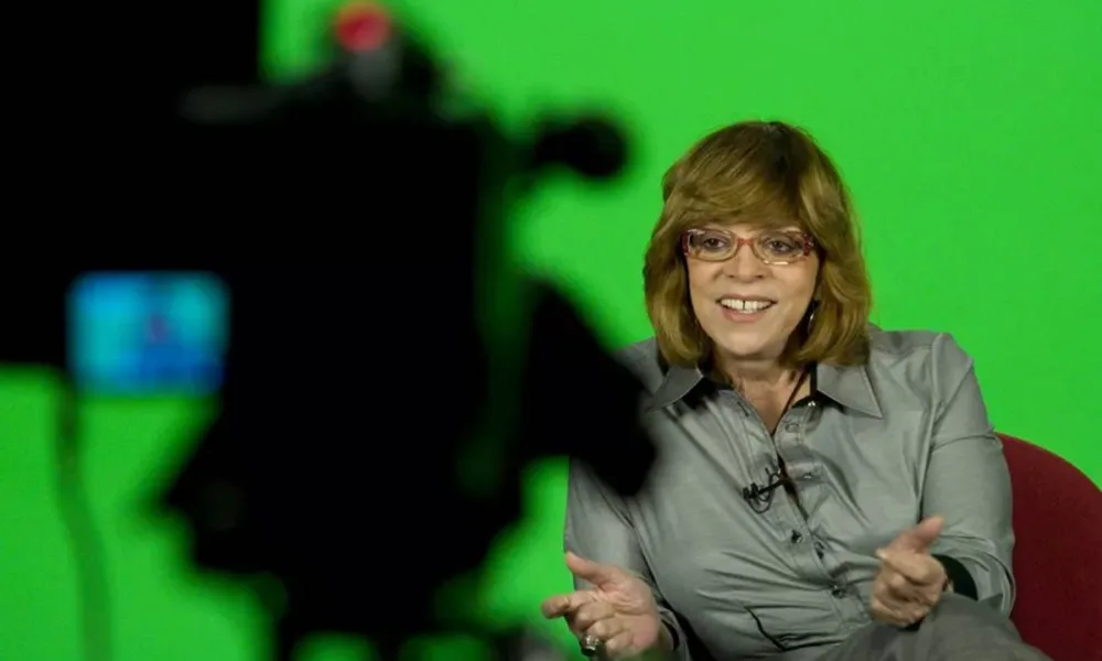 Gloria Perez em 2011 sentada em uma poltrona para dar uma entrevista. Ela sorri e gesticula. Atrás dela há uma tela verde e a sua frente, desfocada, um equipamento de câmera.