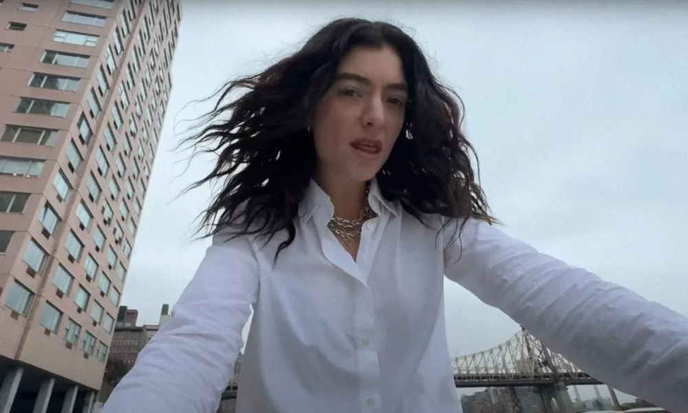 Cantora Lorde com uma camisa branca e colar em corrente de ouro com cabelos ao vento, roupa molhada, e câmera capturando-a sutilmente de um ângulo inferior, no clipe de "What Was That".