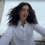 Cantora Lorde com uma camisa branca e colar em corrente de ouro com cabelos ao vento, roupa molhada, e câmera capturando-a sutilmente de um ângulo inferior, no clipe de "What Was That".