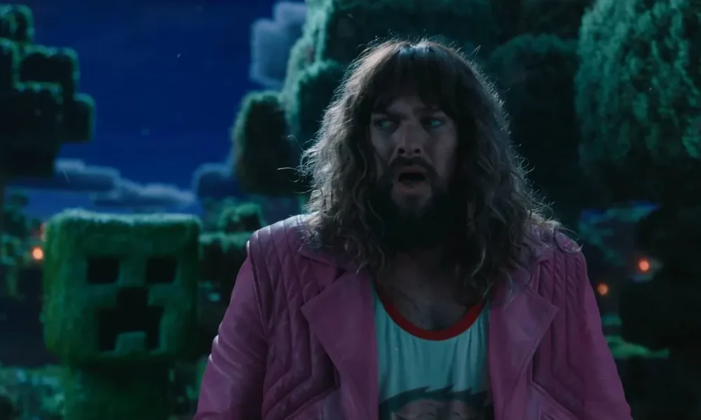 Jason Momoa boquiaberto e com medo interpretando Garrett em "Um Filme Minecraft". Atrás dele há um Creeper, prestes a explodir. Está de noite.
