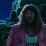 Jason Momoa boquiaberto e com medo interpretando Garrett em "Um Filme Minecraft". Atrás dele há um Creeper, prestes a explodir. Está de noite.