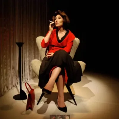 Beth Goulart como Clarice Lispector na peça "Simplesmente Eu, Clarice Lispector", caracterizada como a autora, com um vestido vermelho, saia e sapatos escuros, pernas cruzadas, e segurando um cigarro.