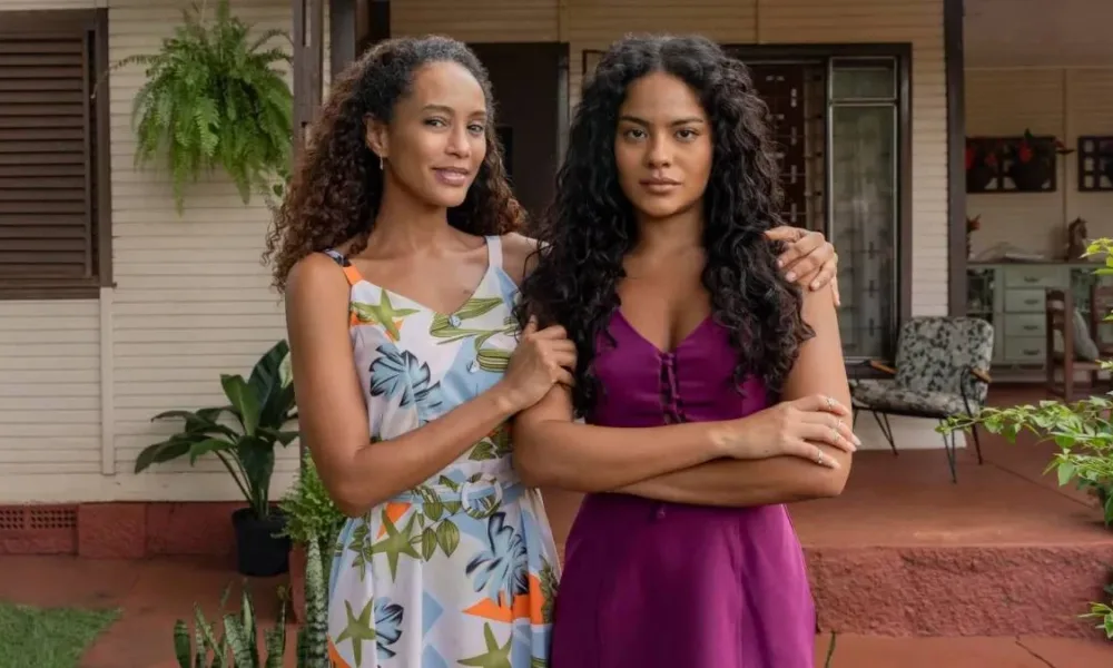 Taís Araújo e Bella Campos como Raquel e Maria de Fátima Acioli, protagonistas do remake de 2025 de "Vale Tudo", posando para uma foto. A mãe abraça a filha, de braços cruzados.