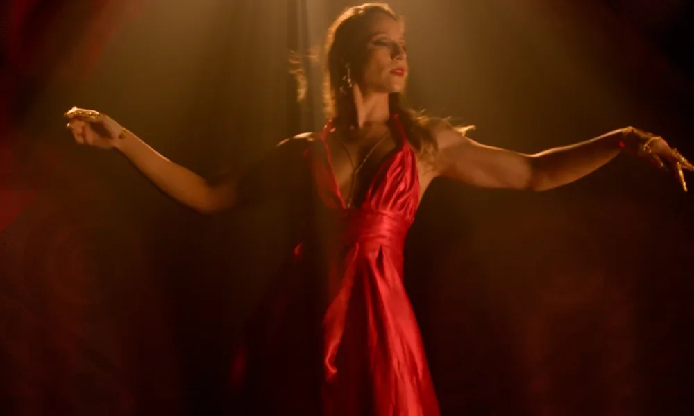 Cartaz do filme "Do Sul, a Vingança", com a protagonista de vestidos vermelhos olhando lateralmente e preparando um passo de dança. Há luz que vem de cima em um fundo de teatro escuro.