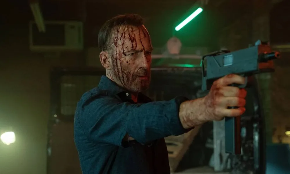 Bob Odenkirk como Hutch Mansell em "Anônimo 2", empunhando uma arma e a apontando com a mão direita, cabeça ensanguentada, mangas de camisa azul arregaçadas. Ele está em um galpão pouco iluminado.