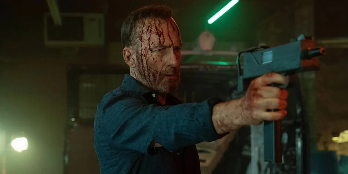 Trailer de Anônimo 2 l Bob Odenkirk Voltando à Ação para Provar Que a Segunda Vez é Melhor Bob Odenkirk como Hutch Mansell em "Anônimo 2", empunhando uma arma e a apontando com a mão direita, cabeça ensanguentada, mangas de camisa azul arregaçadas. Ele está em um galpão pouco iluminado.