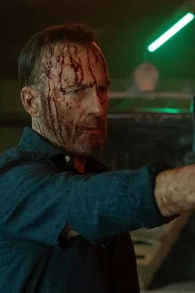 Trailer de Anônimo 2 l Bob Odenkirk Voltando à Ação para Provar Que a Segunda Vez é Melhor Bob Odenkirk como Hutch Mansell em "Anônimo 2", empunhando uma arma e a apontando com a mão direita, cabeça ensanguentada, mangas de camisa azul arregaçadas. Ele está em um galpão pouco iluminado.
