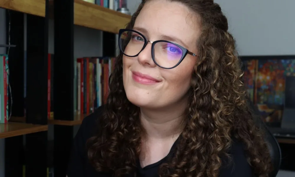 Foto horizontal da autora Pat Müller, sorrindo em quarto com estante ao fundo.