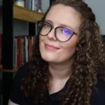 Foto horizontal da autora Pat Müller, sorrindo em quarto com estante ao fundo.