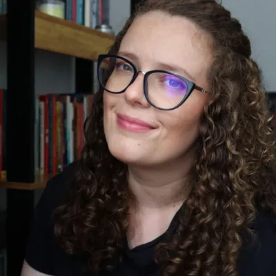 Foto horizontal da autora Pat Müller, sorrindo em quarto com estante ao fundo.