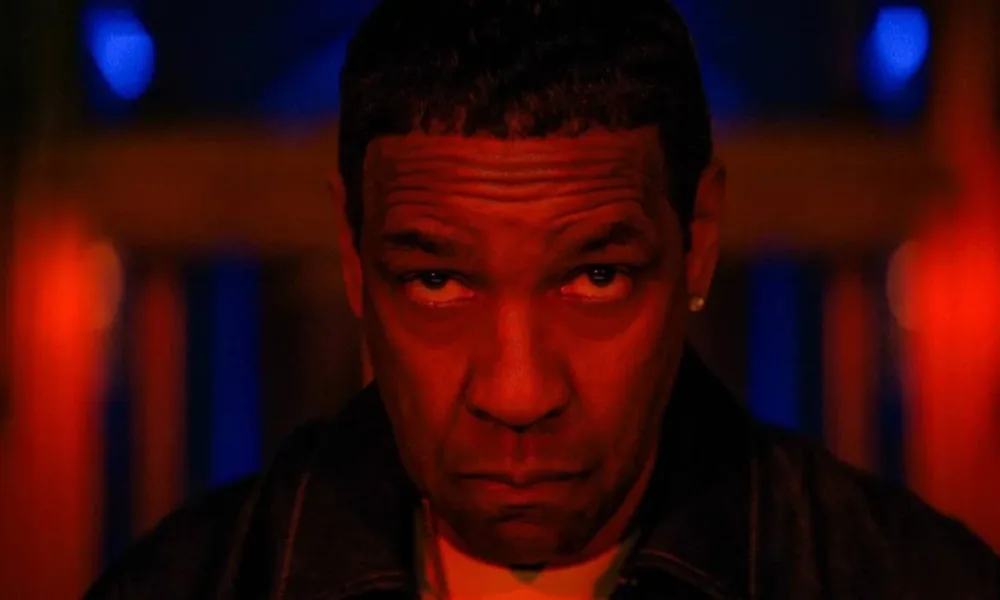 Denzel Washington sob luzes vermelhas, cabeça levemente inclinada para baixo, semblante sério, encarando a câmera centralizado nela como o protagonista do filme de 2025 de Spike Lee "Highest 2 Lowest".