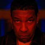 Denzel Washington sob luzes vermelhas, cabeça levemente inclinada para baixo, semblante sério, encarando a câmera centralizado nela como o protagonista do filme de 2025 de Spike Lee "Highest 2 Lowest".