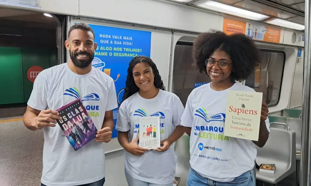 Três pesssoas com camisetas da iniciativa "Embarque na Leitura" em 2025, distribuindo livros no MetrôRio. Cada uma segura um livro, em ordem: "Os clãs da lua", "Mentirosos", e "Sapiens: uma breve história da humanidade".