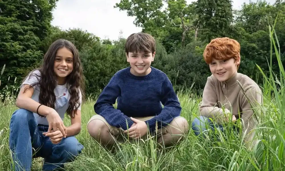 O novo trio de atores que interpretará os protagonistas de Harry Potter, agachados em um gramado em céu diurno: Arabella Stanton (Hermione), Dominic McLaughlin (Harry), e Alastair Stout (Ron).