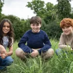 O novo trio de atores que interpretará os protagonistas de Harry Potter, agachados em um gramado em céu diurno: Arabella Stanton (Hermione), Dominic McLaughlin (Harry), e Alastair Stout (Ron).