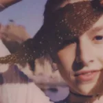 HUnter Schafer interpretando Jules em "Euphoria". Atriz está com braço direito levantado (nossa esquerda da tela), olhando diretamente para a câmera, semi-dobrado, segurando duas folhas douradas pendentes, e um sorriso mostrando parcialmente os dentes de baixo.