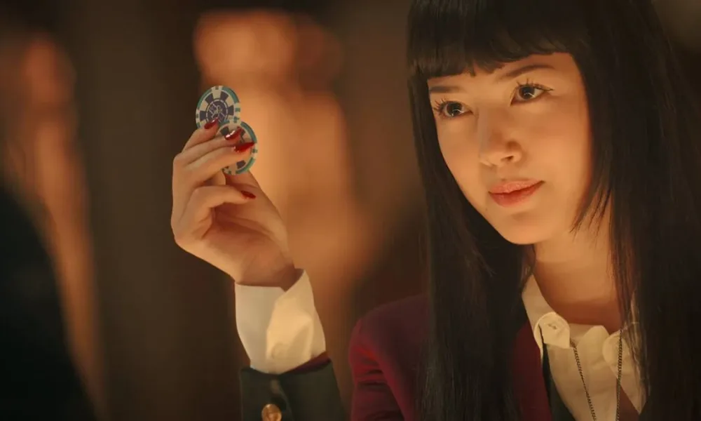 Yumeko de "Kakegurui: Bet" segurando duas fichas e olhando para o personagem do outro lado da tela, com um sorriso levemente malicioso.