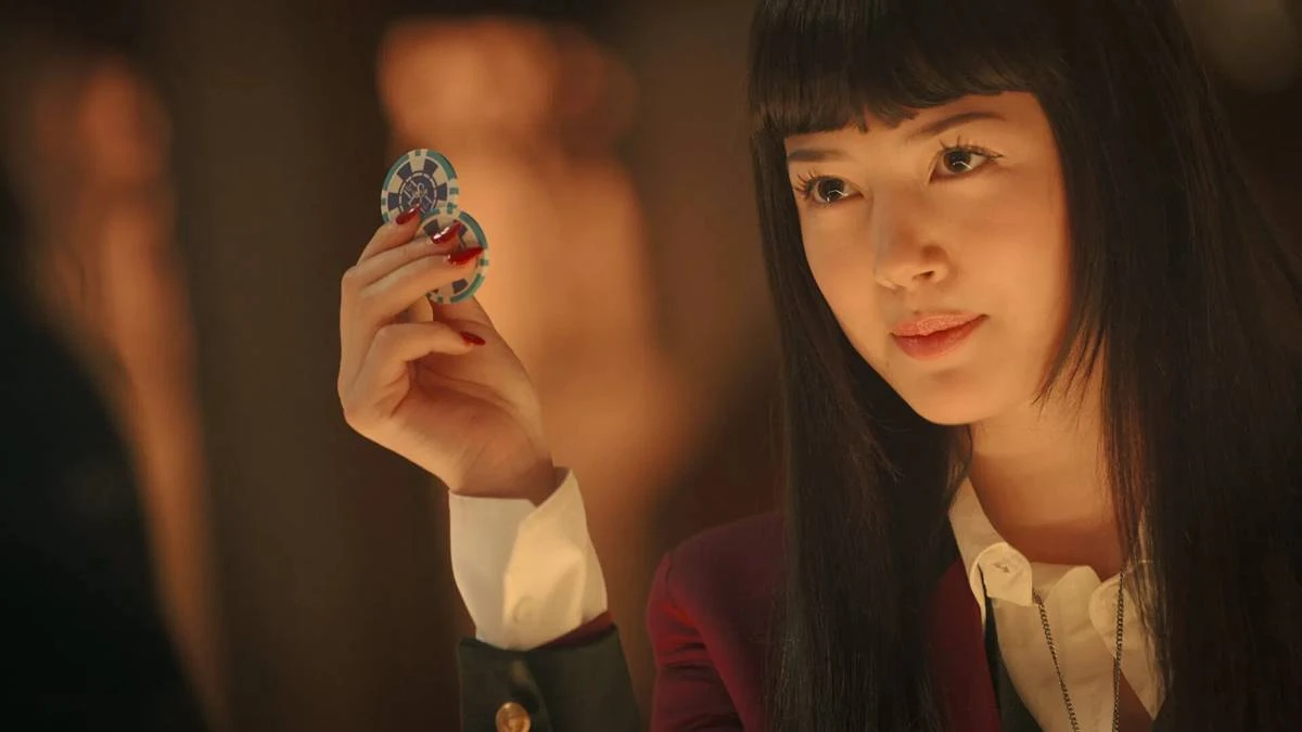 Kakegurui: Bet | Adaptação Plastificada, Mas Mantém Bom Humor Dentro do Possível Para Temporada 1 3 Yumeko de "Kakegurui: Bet" segurando duas fichas e olhando para o personagem do outro lado da tela, com um sorriso levemente malicioso.