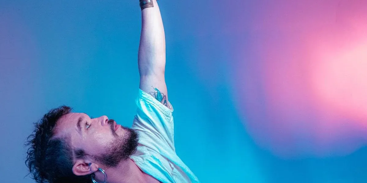 Luas Olivieri, tronco inclinado em arco, fotografado do busto para cima, posando para foto com braço direito erguido. Arista está em um fundo com tons pastéis entre azul e rosa, e escreveu o livro "Patuá e Pé na Terra".