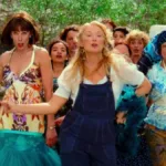 Donna, vestida com uma jardineira, e amigas no primeiro filme de "Mamma Mia", dançando pelas ruas acompanhadas de uma multidão.