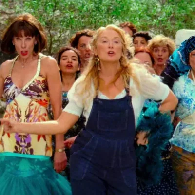 Donna, vestida com uma jardineira, e amigas no primeiro filme de "Mamma Mia", dançando pelas ruas acompanhadas de uma multidão.