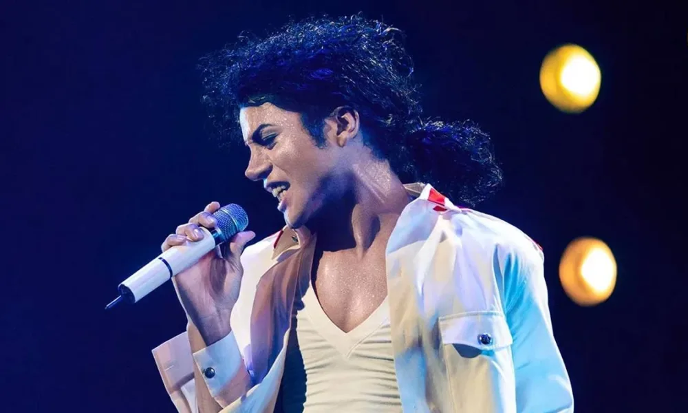 Jaafar Jackson como Michael Jackson na cinebiografia do artista, prevista para 2025, "Michael". O cantor está de camisa e camiseta branca, segurando um microfone branco com a mão direita, inclinado com a cabeça para a direita à esquerda da tela, e cabelo longo preso.