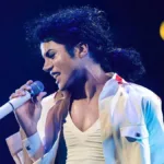 Jaafar Jackson como Michael Jackson na cinebiografia do artista, prevista para 2025, "Michael". O cantor está de camisa e camiseta branca, segurando um microfone branco com a mão direita, inclinado com a cabeça para a direita à esquerda da tela, e cabelo longo preso.