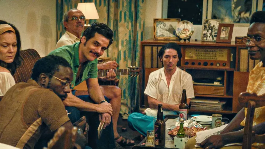 O Agente Secreto | O Baiano Tem o Molho! 1 Wagner Moura como o protagonista de "O Agente Secreto", filme de 2005. Há um senhor a sua esquerda, uma mulher e um homem a sua direita, sentados todos em um sofá e uma poltrona, uma mulher a sua frente sentada na poltrona. Todos se viram para olhar alguém que está atrás da câmera, com a exceção do rapaz, sentado no chão, aéreo.