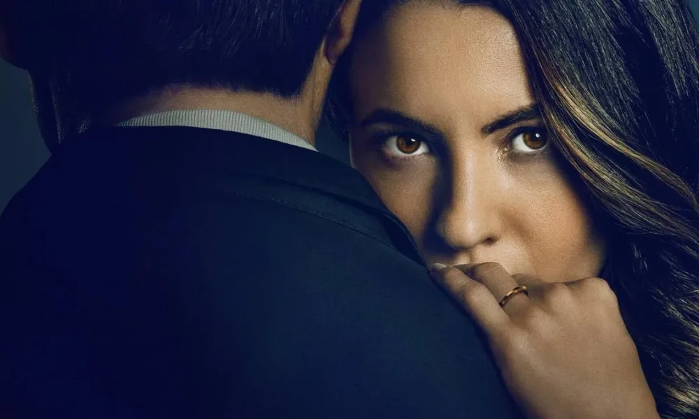 Ivana Baquero, atriz protagonista de "O Jogo da Viúva", em imagem de capa promocional para filme da Netflix. Ela está com o rosto encoberto pelos ombros de um homem, mão esquerda sobre ele, revelando a aliança, e um olhar enigmático. O homem está de terno e de costas.