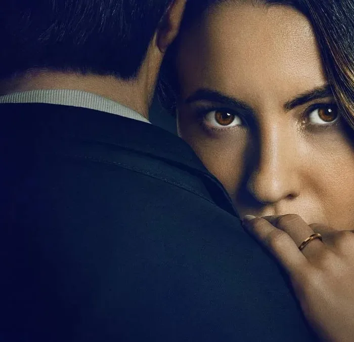 Ivana Baquero, atriz protagonista de "O Jogo da Viúva", em imagem de capa promocional para filme da Netflix. Ela está com o rosto encoberto pelos ombros de um homem, mão esquerda sobre ele, revelando a aliança, e um olhar enigmático. O homem está de terno e de costas.