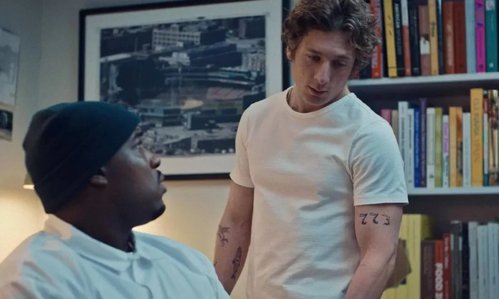 Thumbnail do episódio 7 da terceira temporada de "O Urso", no Disney+ Marcus, corpo virado pela metade para a câmera, encara Carmy, que olha para a mesa em que ele está sentado. Ambos estão vestindo branco.