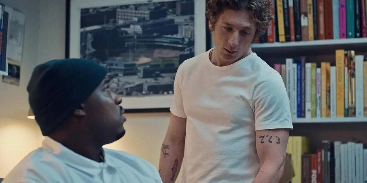 Thumbnail do episódio 7 da terceira temporada de "O Urso", no Disney+ Marcus, corpo virado pela metade para a câmera, encara Carmy, que olha para a mesa em que ele está sentado. Ambos estão vestindo branco.