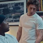 Thumbnail do episódio 7 da terceira temporada de "O Urso", no Disney+ Marcus, corpo virado pela metade para a câmera, encara Carmy, que olha para a mesa em que ele está sentado. Ambos estão vestindo branco.