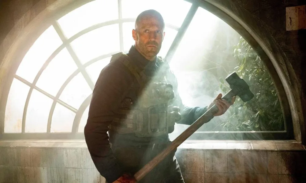 Jason Statham como Levon Cade, protagonista de "Resgate Implacável". Ele segura com as duas mãos uma grande marreta e está vestido com um colete. De cara séria, ele está de frente para câmera e de trás para uma janela em semicírculo com luz natural diurna passando pelo ambiente escuro.